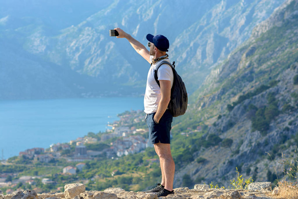 A man with a backpack pictures himself against the backdrop of the mountains and the old town of Kotor in Montenegro. - Foto, afbeelding