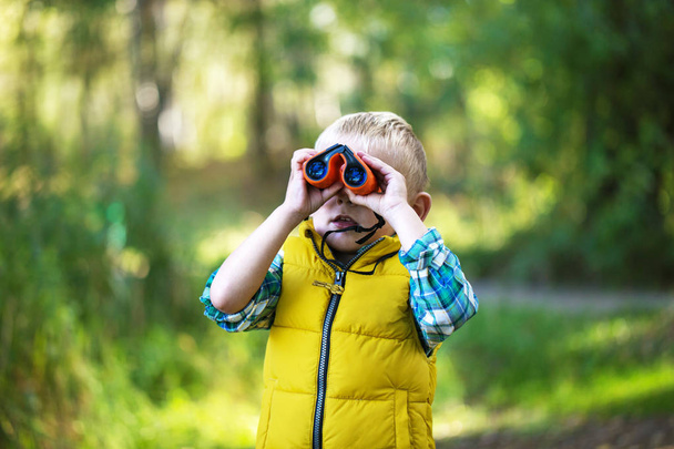 The little boy young researcher looks up and exploring with binoculars environment in the green forest on a sunny day - Fotó, kép