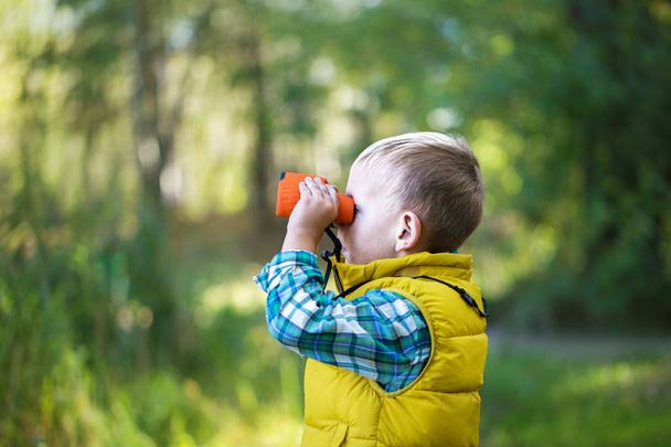 The little boy young researcher looks up and exploring with binoculars environment in the green forest on a sunny day - Foto, imagen