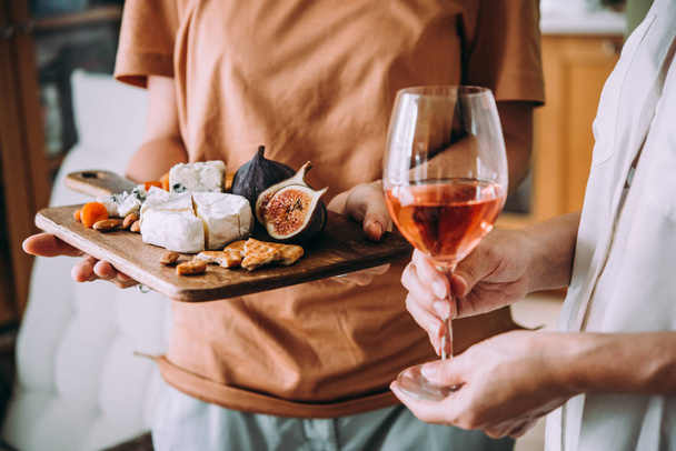 Hands holding a glass of wine and a wooden board with different kinds of cheese and dried fruits. Dinner or aperitivo party concept. - Фото, изображение