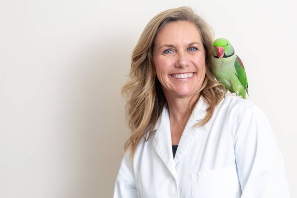 Щаслива і доброзичлива жінка-ветеринар з зеленим птахом Олександріна на плечі
 - Фото, зображення