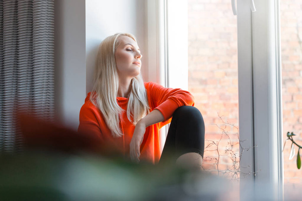 Blond young woman wearing sport clothes resting by the window at home.  - Valokuva, kuva