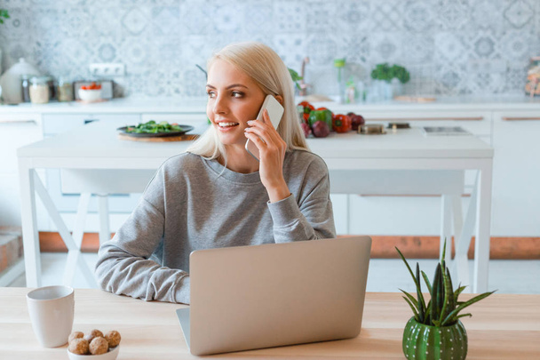 Blonde young woman sitting at the table in the kitchen and talking on smart phone. Laptop on the table.  - Фото, изображение