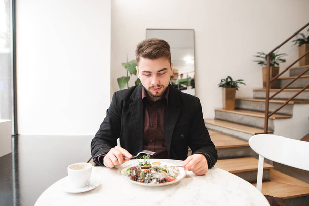 Άνθρωπος των επιχειρήσεων με μια γενειάδα κάθεται σε ένα άνετο εστιατόριο στο τραπέζι, τρώγοντας σαλάτα και εξετάζοντας τη φωτογραφική μηχανή. Πορτρέτο ενός επιχειρηματία στο πρωινό στο φωτεινό ΚΠΑ. - Φωτογραφία, εικόνα