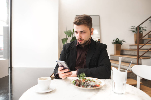 Hombre de negocios con barba sentado en un acogedor restaurante a la mesa, comiendo ensalada y mirando a la cámara. Retrato de un hombre de negocios en el desayuno en el café brillante
. - Foto, Imagen