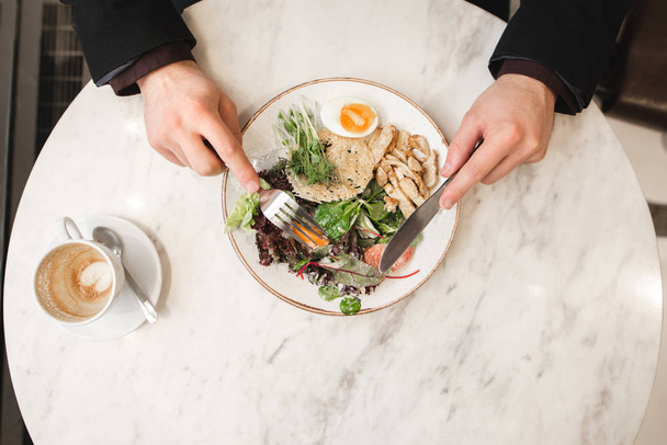 Man eats an appetizing salad with meat with a fork and a knife in his hands, close up, top view, cup of coffee on a table. Business man dining at a restaurant. Lunch concept - Photo, Image