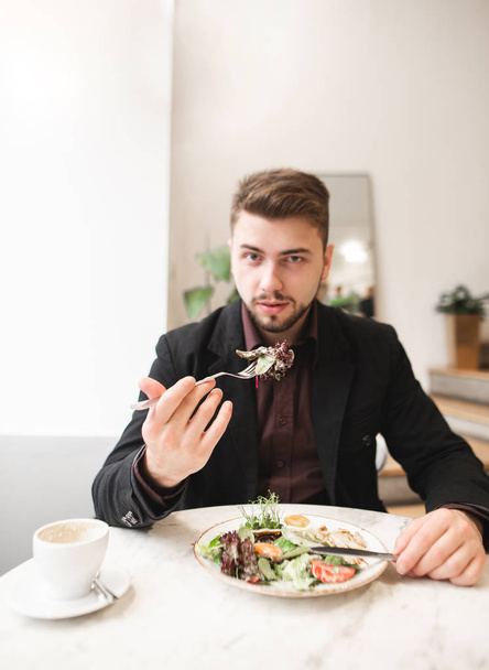 Przystojny mężczyzna siedzi z przytulną restaurację światła i zjada płyta sałatka i pije kawę. Działalności człowieka dania dobre jedzenie w restauracji, Sałatka i patrzy w kamerę. - Zdjęcie, obraz