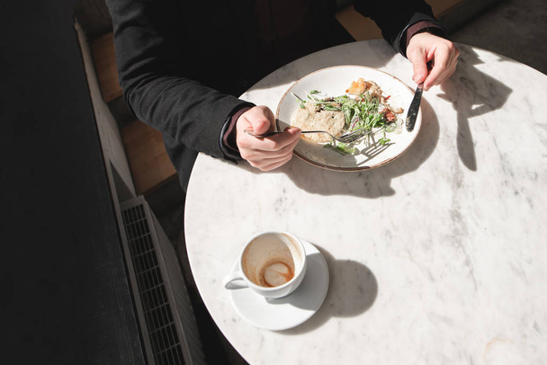 Kopje koffie en een plaat van salade op tafel. Man eet een salade met een vork en een mes in zijn handen. Bovenaanzicht. Gezonde voeding. Lunchpauze. Licht en schaduwen. Copyspace - Foto, afbeelding