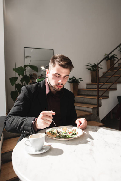 Портрет делового человека, сидящего в уютном ресторане и обедающего с салатом и кофе. Человек в деловом костюме ест в легком ресторане
. - Фото, изображение