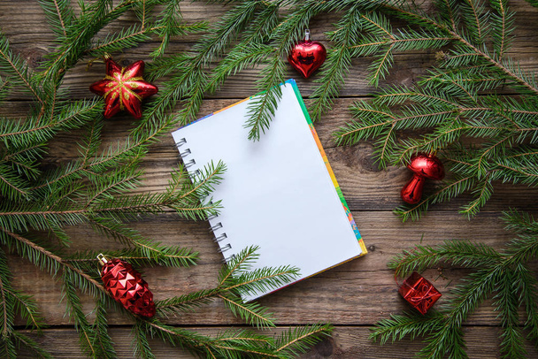 Notebook, white sheet of paper in a frame of Christmas tree branches with red toys on a wooden rustic background, top view, with copy space - Foto, Imagem
