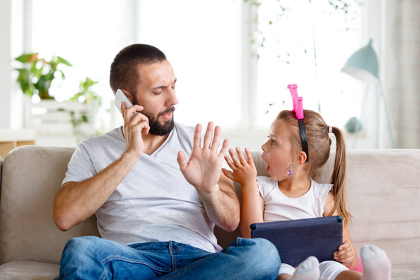 Young father sitting on sofa in living room and talking on phone while his litte daughter disturbing him.  - Fotó, kép