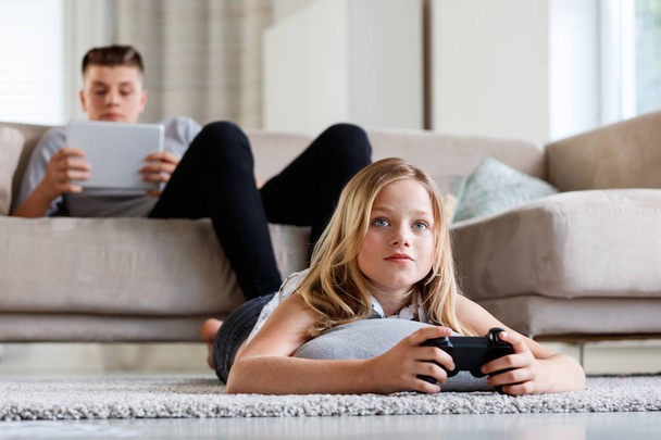 Cute girl lying on stomach on carpet and playing video game while her brother sitting on sofa and using a digital tablet.  - 写真・画像