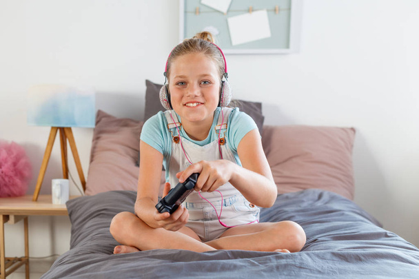 Smiling girl wearing headphone, sitting on bed in her room, holding controller and playing video game. - Photo, image