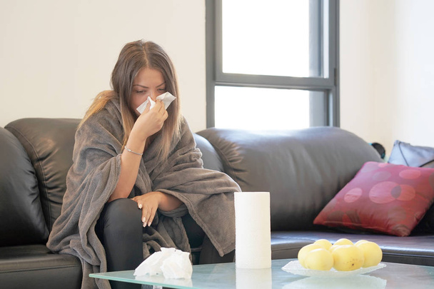 Attractive woman feeling cold, freezing, with napkin in a hand, wrapped in blanket, sitting on the sofa. Unhappy upset tired woman is sitting on a sofa at home suffering from a cold and using napkins. - Photo, Image