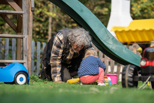 Dziadek, grając z jego wnuczka poza w podwórku, jak siedzieć na trawie pod zjeżdżalnią i grać z piasku zabawki i wiadra.   - Zdjęcie, obraz