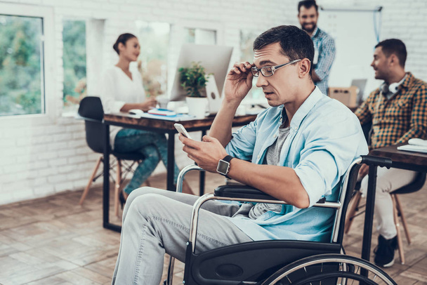 Man in Glasses on Wheelchair with Phone in Office. Talking on Phone. Disabled Young Man. Man on Wheelchair. Recovery and Healthcare Concepts. Teamwork in Office. Young Worker. Sitting Man. - 写真・画像