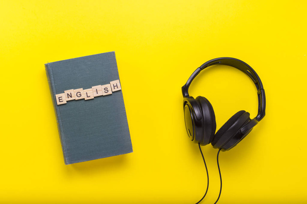 Βιβλίο με μπλε εξώφυλλο με κείμενο Ελληνικά και μαύρα ακουστικά σε κίτρινο φόντο. Έννοια της ακουστικά βιβλία, αυτο-εκπαίδευση και εκμάθηση της αγγλικής γλώσσας κατά τρόπο ανεξάρτητο. Επίπεδη lay, κορυφαία προβολή. - Φωτογραφία, εικόνα