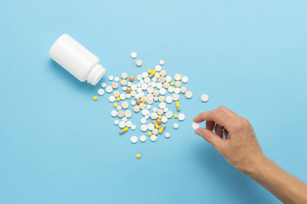 Female hand takes a pill from a pile of scattered pills and a white bottle on a blue background. Concept of taking drugs in case of illness, pharmacetiya, medicine. Flat lay, top view. - Photo, Image