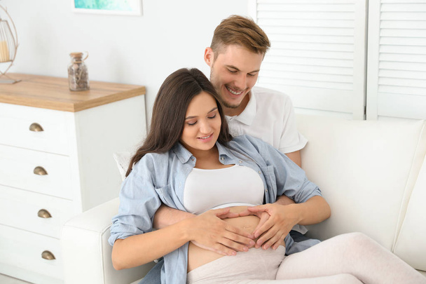 Young husband and his pregnant wife resting on sofa at home - Fotoğraf, Görsel