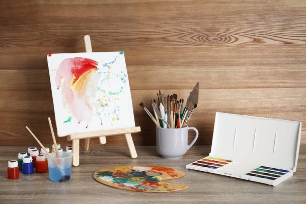 Easel with abstract painting and set of professional art supplies on table against wooden background - Fotoğraf, Görsel