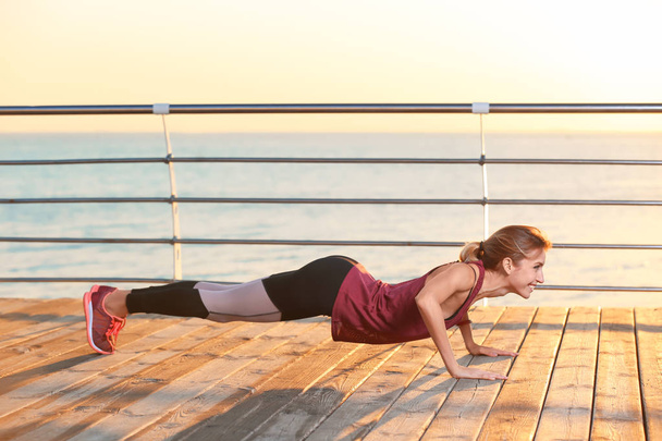 Young woman doing fitness exercises on pier in morning - Фото, изображение