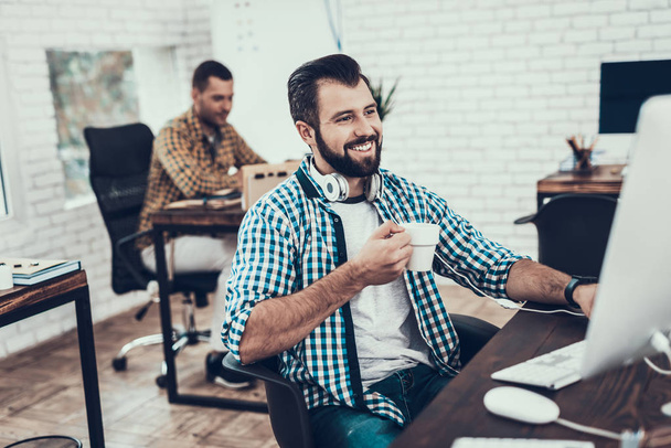 Smiling Bearded Man Sitting on Chair in Office. Didital Device on Desk. Modern Office, Black Chair. Man with Unshaved Face. Work in Office Concept. Young Worker. Man in White Room. White Gadget. - Фото, изображение