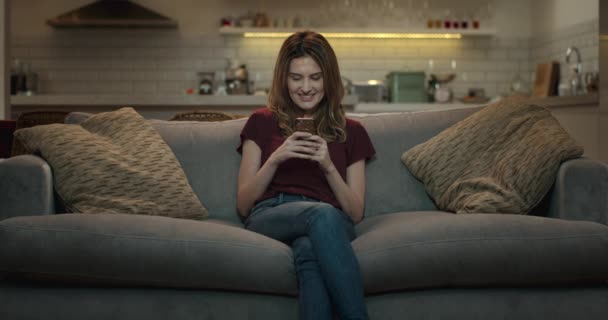 Frau sitzt auf einem Sofa und textet mit ihrem Smartphone mit einem Freund. - Filmmaterial, Video