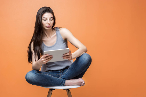 Portrait of a young beautiful girl with a tablet in her hands sitting in the chair against orange background. Lifestyle, people and technology concept - Fotografie, Obrázek