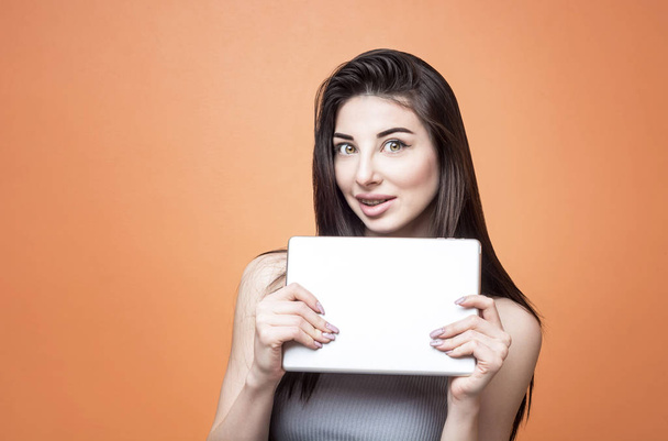 Closeup portrait of a young excited brunette girl with tablet in her hands looking at the camera with open mouth against orange background. Lifestyle, people and technology concept - Фото, изображение
