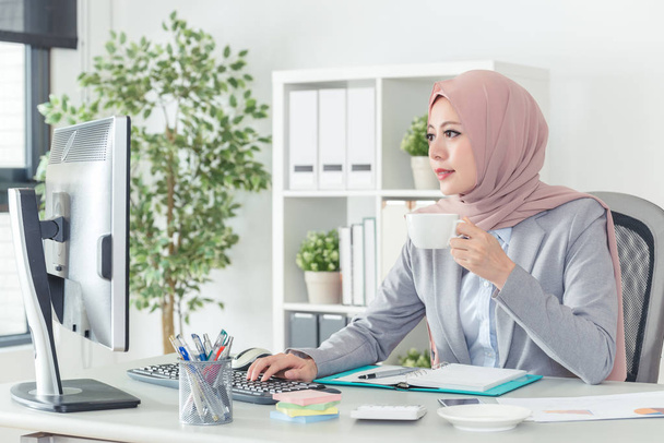 successful Muslim office lady sitting in her office desk and holding a cup of coffee. Portrait of a confident businesswoman working on computer while drinking a coffee cup in the morning. - Foto, imagen