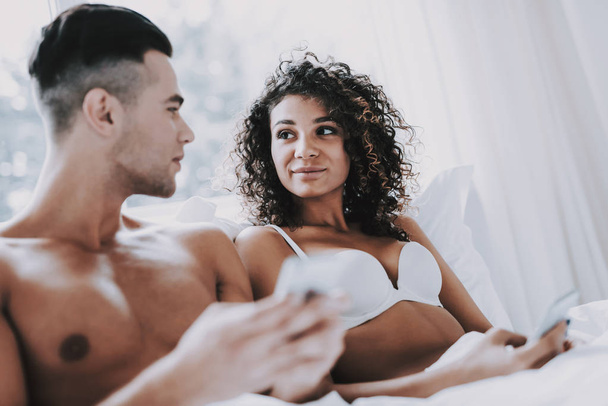 Young Beautiful Couple Using Smartphones in Bed. Attractive Woman and Handsome Man Lying in Bed Together and Using Cellphones. Happy Smiling Couple in Underwear Relaxing in Bed at Home. - Фото, изображение