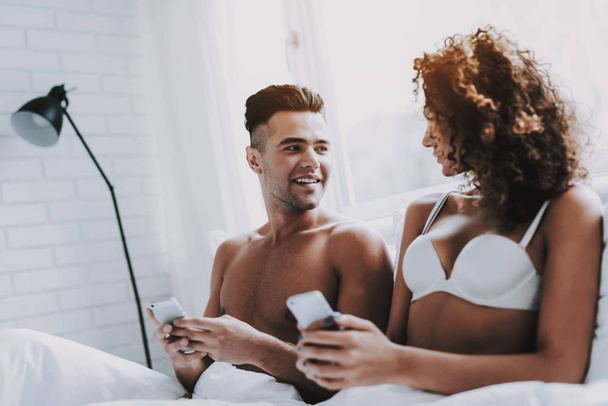 Young Beautiful Couple Using Smartphones in Bed. Attractive Woman and Handsome Man Lying in Bed Together and Using Cellphones. Happy Smiling Couple in Underwear Relaxing in Bed at Home. - Photo, image