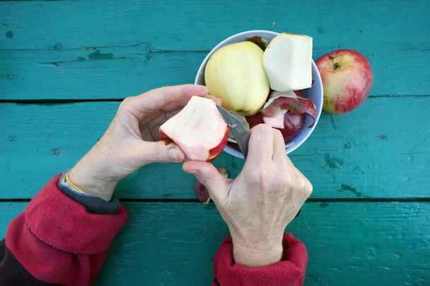 The elderly gardener  woman cleans the skins from  apples to eat fruits . Green wwoden table as background - Photo, Image