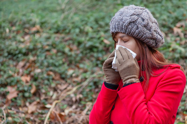 Fiatal nő szenved a megfázás vagy influenza, fúj az orrát, a fehér papír zsebkendőt visel egy piros kabátot, egy sapka és kesztyű téli erdő  - Fotó, kép