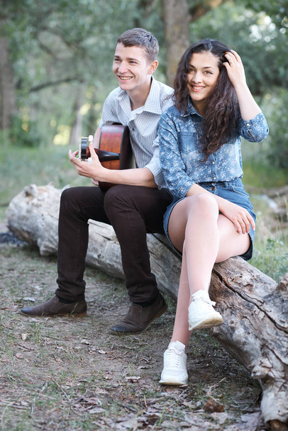young couple sitting on a log in the forest and playing guitar, summer nature, romantic feelings - Photo, Image