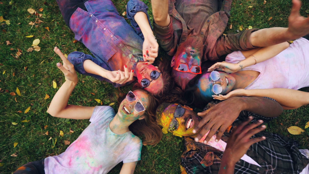 Widok z góry szczęśliwy przyjaciół płci męskiej i żeńskiej, leżąc na trawie w parku z twarze i ubrania pokryte farbą jasne, patrząc na aparatu i ruchu rąk, uśmiechnięty. - Materiał filmowy, wideo
