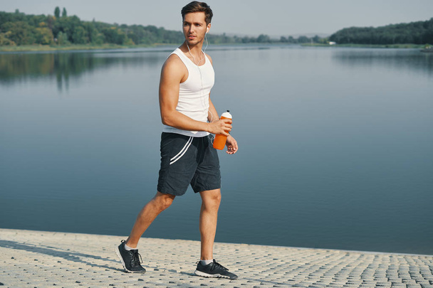 Портрет улыбающегося и расслабляющего спортсмена с бутылкой воды в руках. Спортсмен в черной спортивной одежде в белых наушниках наслаждается утром, когда бегает трусцой возле озера по соседству
 - Фото, изображение