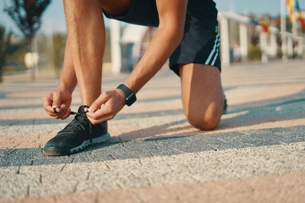 Sportsman ties his black sneakers. Athletic man tying shoelaces while getting ready to run. Smart watch on left hand. Closeup shot - Zdjęcie, obraz
