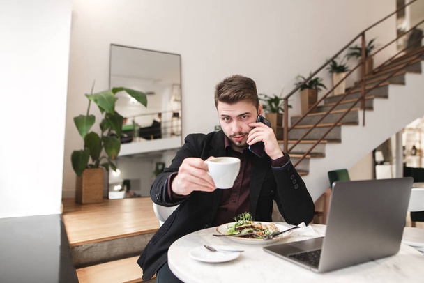 L'homme barbu en costume se trouve dans un café avec une assiette de salade et un ordinateur portable et offre une tasse de café. Homme occupé dans le café montre une tasse de café dans la caméra
. - Photo, image
