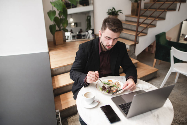 Πορτραίτο ενός ανθρώπου που κάθεται σε ένα ζεστό εστιατόριο με ένα φορητό υπολογιστή και ένα smartphone στο τραπέζι και τρώει μια σαλάτα. Πολυάσχολος άνθρωπος πιάτα σε ένα καφέ. - Φωτογραφία, εικόνα