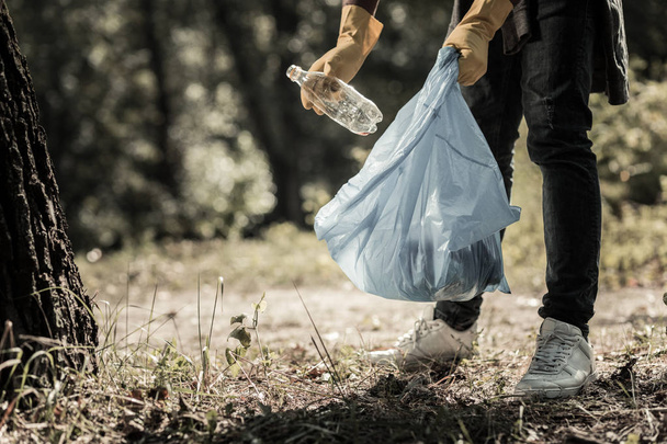 Joven alumno con jeans y zapatillas de deporte recogiendo botellas vacías en el bosque
 - Foto, Imagen