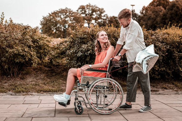 Kind man looking at his disabled girlfriend while pushing her wheelchair - Photo, Image