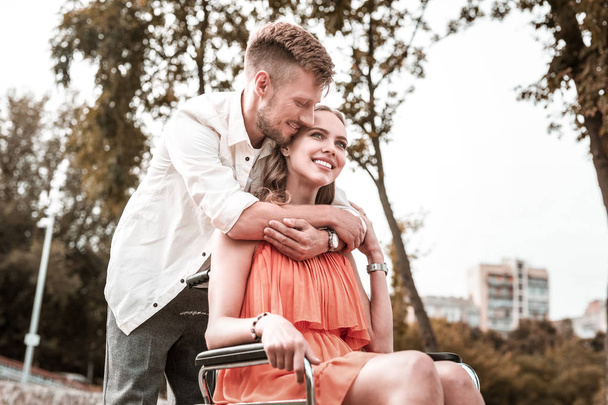 Calm man smiling and hugging girlfriend in wheelchair - Zdjęcie, obraz
