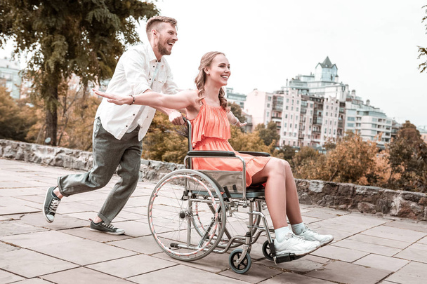 Emocional hombre empujando la silla de ruedas y su novia discapacitada sonriendo
 - Foto, Imagen