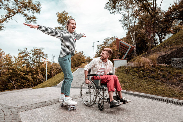 Positive man sitting in wheelchair and his girlfriend riding her skateboard - Foto, afbeelding