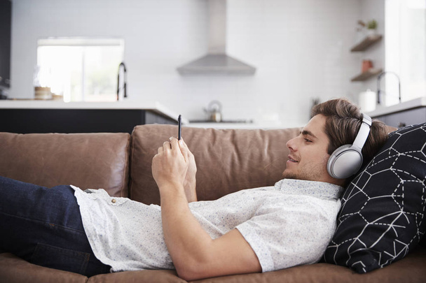 Man Wearing Wireless Headphones Lying On Sofa At Home Streaming From Mobile Phone - Foto, immagini