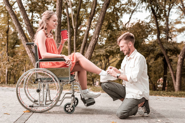 Handsome man looking glad while tying shoelaces of disabled girlfriend - Foto, afbeelding