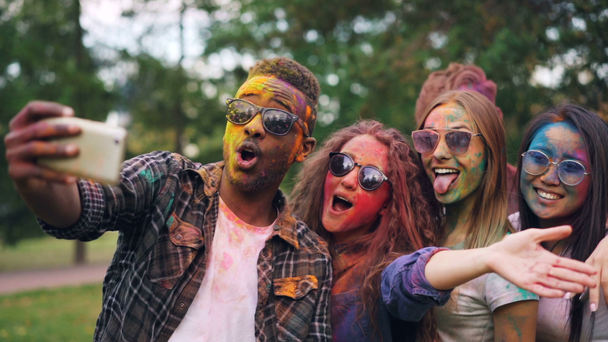 Excited friends girls and guys are taking selfie with colored faces and hair using smartphone, stylish young man is jumping and laughing. Modern technology and party concept. - Footage, Video