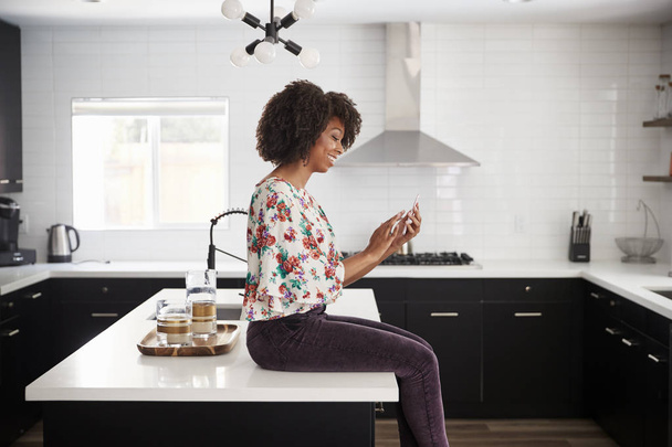 Woman At Home Sitting On Kitchen Island Whilst Using Mobile Phone - Foto, Bild