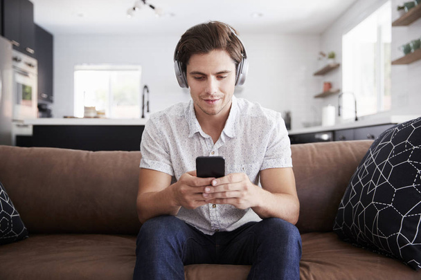 Man Wearing Wireless Headphones Sitting On Sofa At Home Streaming From Mobile Phone - Foto, Imagem